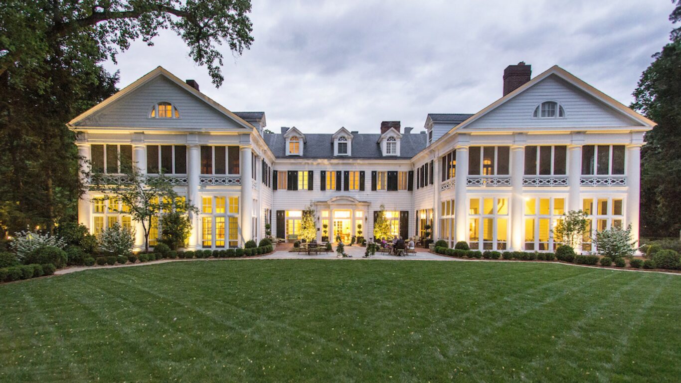 Stately white residence at dusk with warmly lit windows and a wide lawn